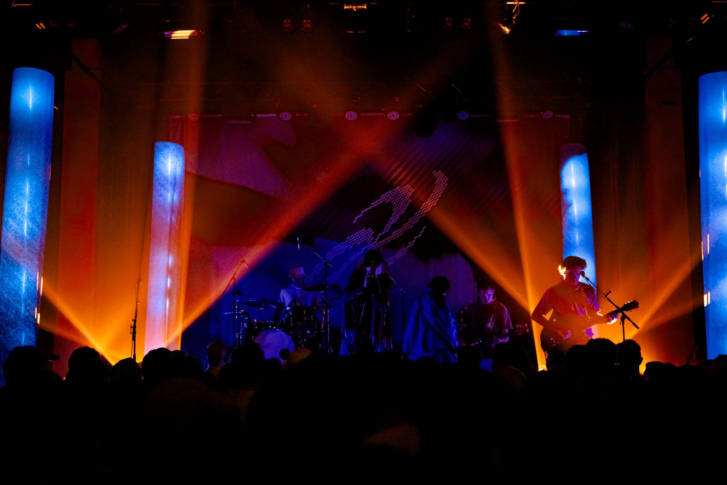 Warm orange lights light up a stage from each corner, with beams of blue cutting up through them
